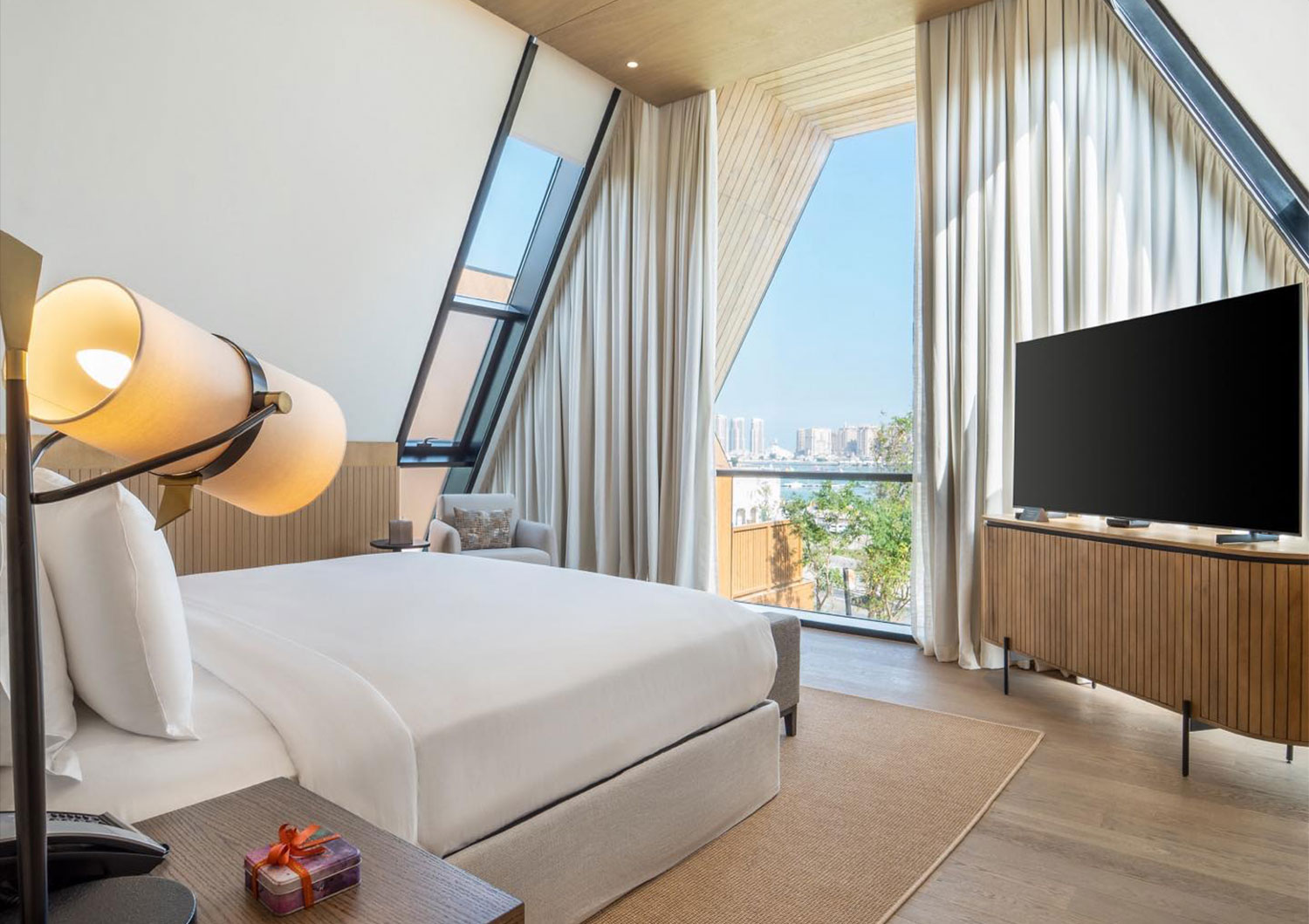 Bedroom in a Katara Hills villa, Doha: white upholstered bed under a triangular glazed roof, vertical timber batten wall and a fluted timber TV unit on metal legs