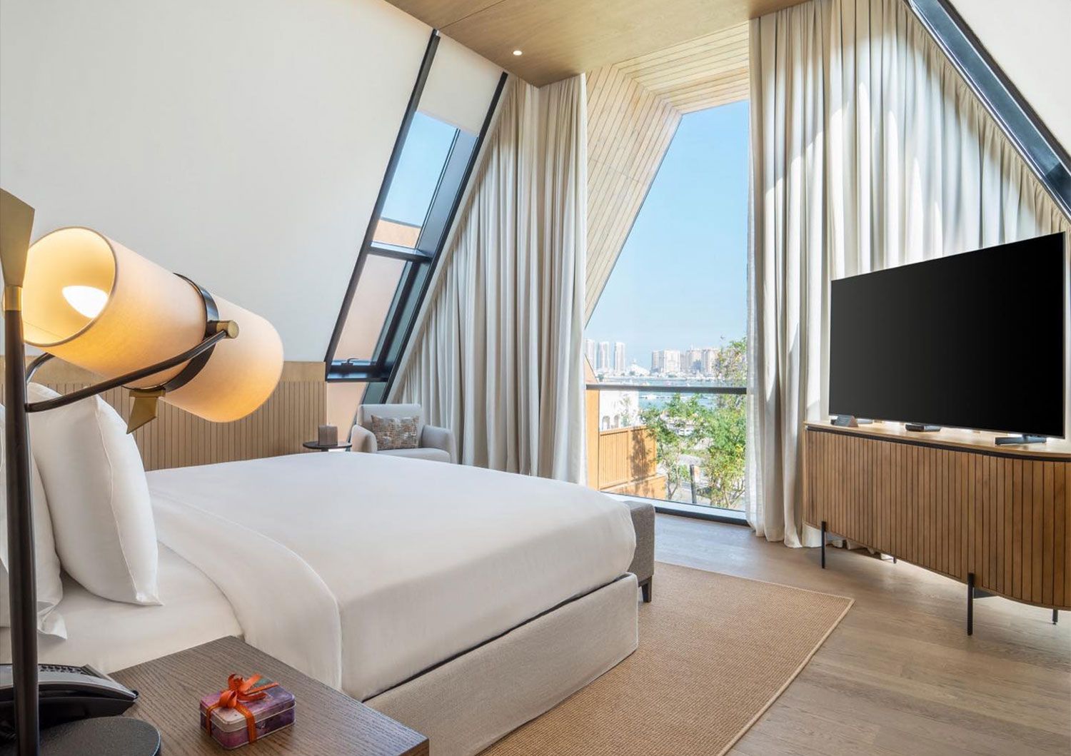 Katara Hills villasında yatak odası: beyaz nevresimli yatak, ahşap kaplamalı TV ünitesi ve üçgen cam cephe